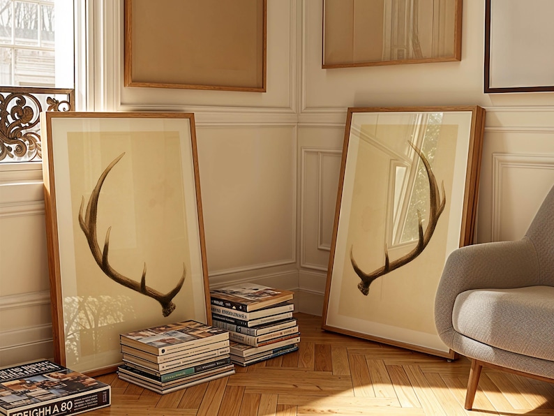 May include: Two framed prints of a brown antler on a beige background. The prints are hanging on a white wall in a room with wooden floors. There are books stacked on the floor in front of the prints.