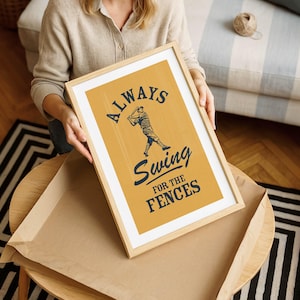 May include: A framed print with the text "ALWAYS Swing FOR THE FENCES" in navy blue over a baseball player illustration on a mustard yellow background. The print is in a light wood frame.