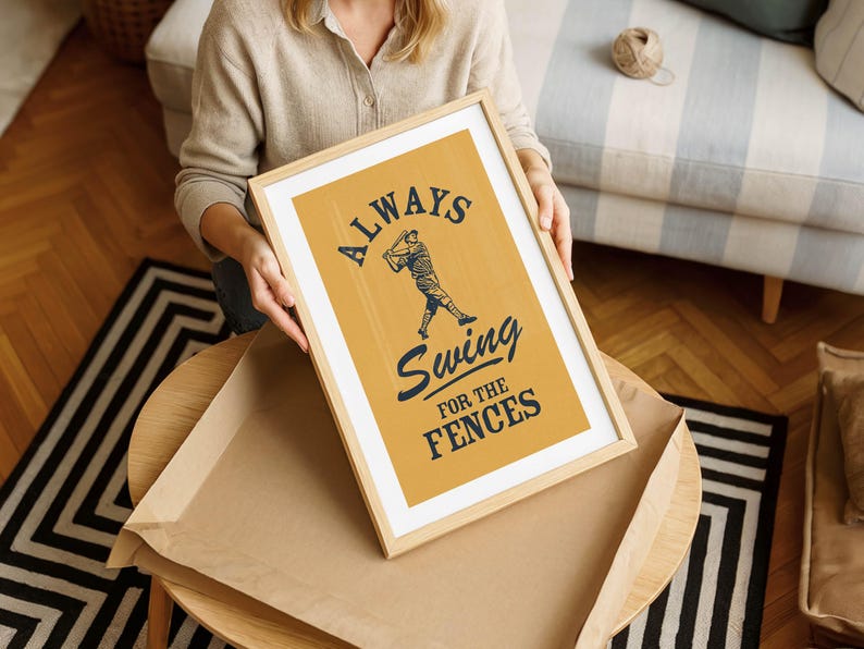 May include: A framed print with the text "ALWAYS Swing FOR THE FENCES" in navy blue over a baseball player illustration on a mustard yellow background. The print is in a light wood frame.