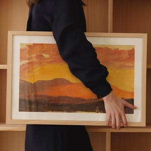 May include: A framed landscape painting with a warm color palette of orange, yellow, and brown. The artwork depicts a sunset over a mountain range. The frame is made of light-colored wood.