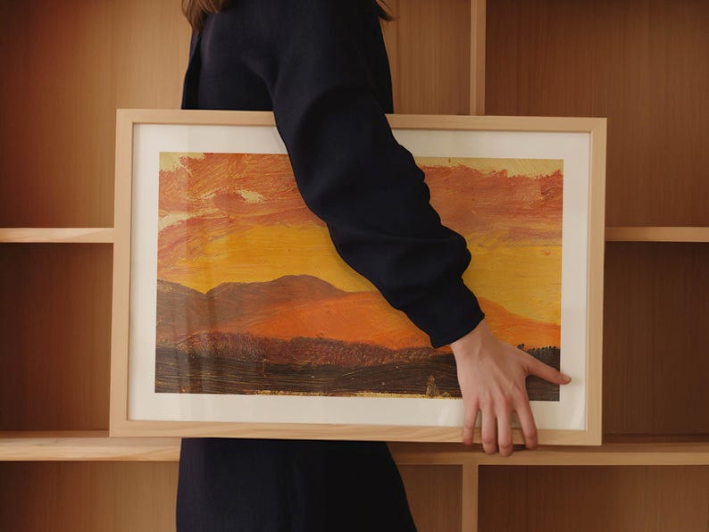 May include: A framed landscape painting with a warm color palette of orange, yellow, and brown. The artwork depicts a sunset over a mountain range. The frame is made of light-colored wood.