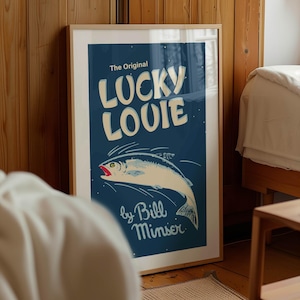 May include: Framed art print featuring a vintage-style illustration of a fish with the text "LUCKY LOUIE" in large, white, bold letters. The artwork is set against a dark blue background, with the artist's name at the bottom.