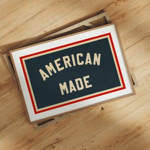 May include: A framed print with the text "AMERICAN MADE" in a vintage font. The text is on a dark blue background, framed by a red and white border, and set in a light brown frame. The print is displayed on a wooden surface.