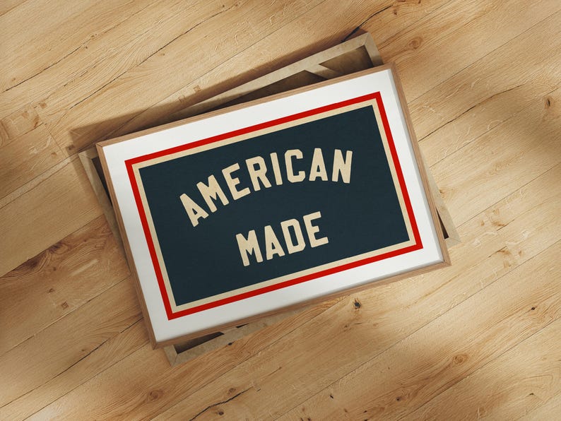 May include: A framed print with the text "AMERICAN MADE" in a vintage font. The text is on a dark blue background, framed by a red and white border, and set in a light brown frame. The print is displayed on a wooden surface.