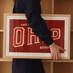 K&ouml;nnte beinhalten: Ein gerahmter Druck mit rotem Hintergrund und wei&szlig;em Text, der "SINCE 1870 OHIO BUCKKEYE" lautet. Der Druck wird von einer Person in einem schwarzen Shirt gehalten.