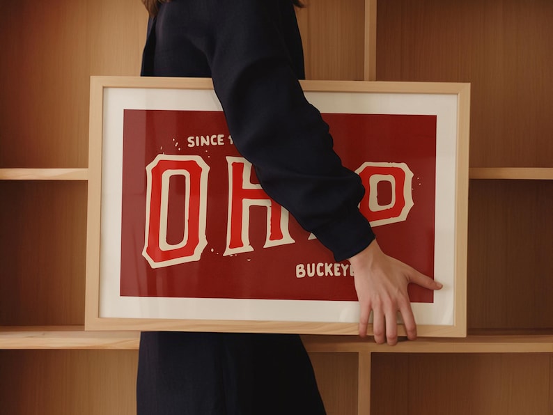 K&ouml;nnte beinhalten: Ein gerahmter Druck mit rotem Hintergrund und wei&szlig;em Text, der "SINCE 1870 OHIO BUCKKEYE" lautet. Der Druck wird von einer Person in einem schwarzen Shirt gehalten.