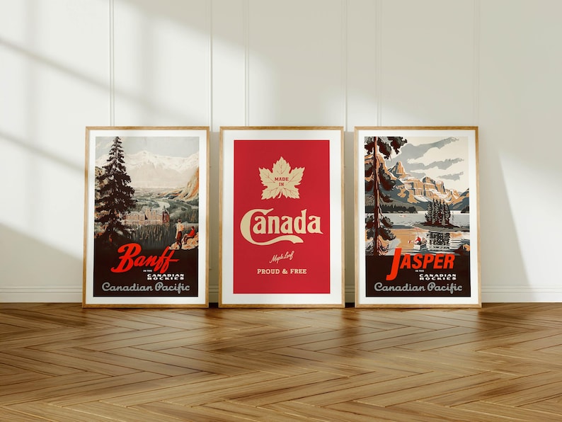 Op de afbeelding: Drie ingelijste vintage reisposters. De posters tonen schilderachtige uitzichten op Banff en Jasper, Canada, en een rode poster met het woord "Canada" en een esdoornblad. De posters zitten in houten frames en worden op een houten vloer getoond.