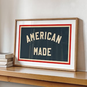 May include: A framed print displaying the text "AMERICAN MADE" in cream-colored lettering on a dark blue background. It features a red and white border, encased in a light brown wooden frame. The print is displayed on a wooden shelf.