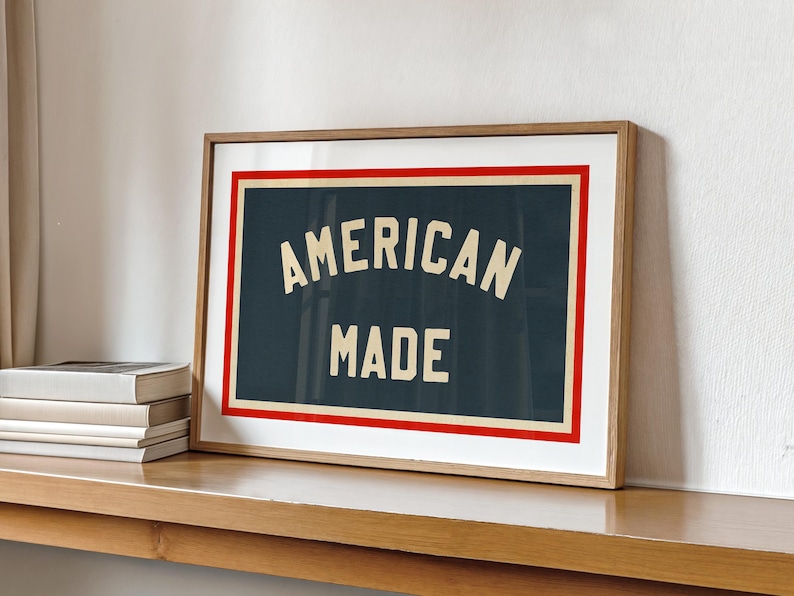 May include: A framed print displaying the text "AMERICAN MADE" in cream-colored lettering on a dark blue background. It features a red and white border, encased in a light brown wooden frame. The print is displayed on a wooden shelf.