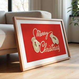 May include: A framed Christmas art print with a red background. The artwork features the words "Merry Christmas" in cursive script, along with two mittens decorated with holly. The frame is light-colored wood.