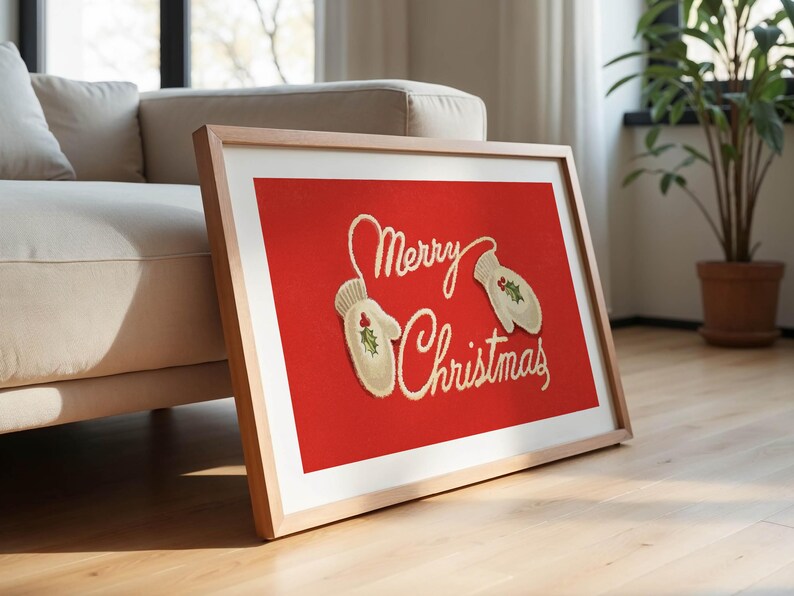 May include: A framed Christmas art print with a red background. The artwork features the words "Merry Christmas" in cursive script, along with two mittens decorated with holly. The frame is light-colored wood.