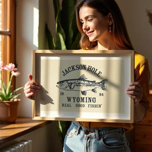 May include: A framed print with a beige background featuring a black illustration of a fish and the words "Jackson Hole Wyoming Real Good Fishin'". The print is held by a person in front of a window.