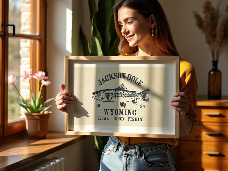 May include: A framed print with a beige background featuring a black illustration of a fish and the words "Jackson Hole Wyoming Real Good Fishin'". The print is held by a person in front of a window.