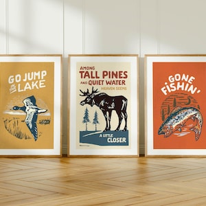 May include: Three framed vintage-style prints with outdoor themes. The prints feature a duck, a moose, and a fish, with text that reads "Go Jump in the Lake", "Among Tall Pines and Quiet Water", and "Gone Fishin'", respectively. The frames are a light brown color.