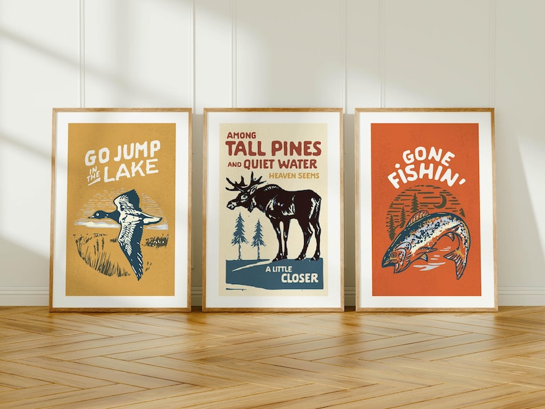 May include: Three framed vintage-style prints with outdoor themes. The prints feature a duck, a moose, and a fish, with text that reads "Go Jump in the Lake", "Among Tall Pines and Quiet Water", and "Gone Fishin'", respectively. The frames are a light brown color.