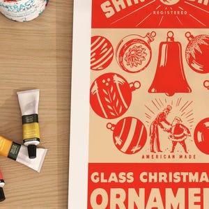 Op de afbeelding: Vintage advertentie voor Shinybrite glazen kerstboomversieringen. De advertentie toont een rode bel en verschillende rode en witte kerstballen met een feestelijk ontwerp. De tekst luidt "Shinybrite Registered American Made Glass Christmas Tree Ornaments".
