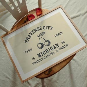 May include: A framed print with a light beige background featuring a vintage-style design. The text reads "Traverse City Michigan Cherry Capitol of the World" with an illustration of cherries. The frame is made of light brown wood.