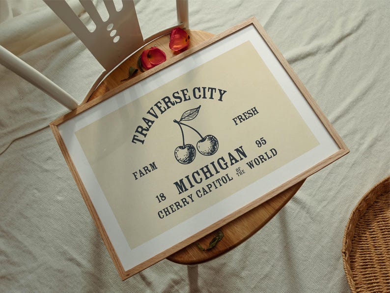 May include: A framed print with a light beige background featuring a vintage-style design. The text reads "Traverse City Michigan Cherry Capitol of the World" with an illustration of cherries. The frame is made of light brown wood.