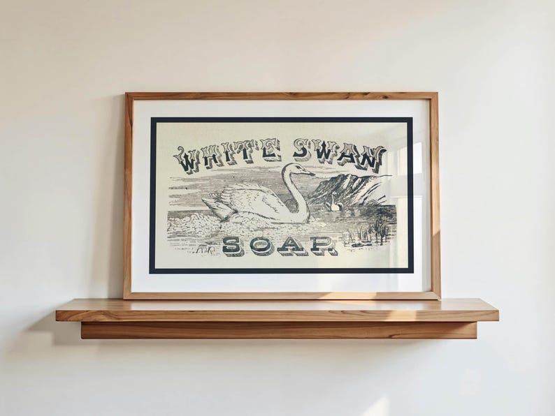 May include: Framed vintage print of a swan with the text "WHITE SWAN SOAP". The artwork is in a wooden frame with a white mount and a dark blue border. The print is displayed on a wooden shelf against a white wall.