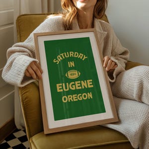 K&ouml;nnte beinhalten: Gerahmtes Poster mit gr&uuml;nem Hintergrund und goldenem Text "Saturday in Eugene Oregon 1894". Das Poster zeigt eine Football-Grafik. Der Rahmen ist hellbraun.