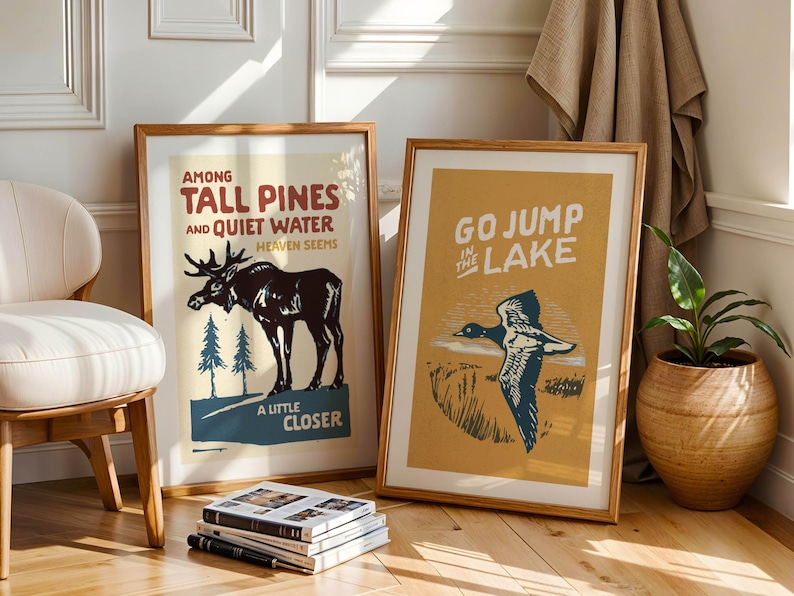 May include: Two framed vintage-style posters with nature scenes. The left poster features a moose in a forest with the text "Among Tall Pines and Quiet Water, Heaven Seems A Little Closer". The right poster features a duck flying over a lake with the text "Go Jump in the Lake".