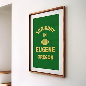 K&ouml;nnte beinhalten: Gerahmtes Poster mit gr&uuml;nem Hintergrund und goldenem Text. Der Text lautet "SATURDAY IN 1894 EUGENE OREGON" mit einer Football-Grafik. Der Rahmen hat eine dunkelbraune Holzfarbe.