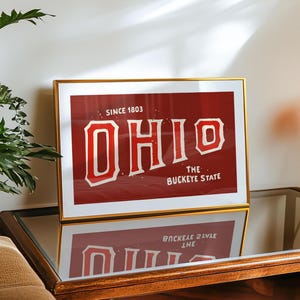 K&ouml;nnte beinhalten: Ein goldfarbener gerahmter Druck mit rotem Hintergrund und wei&szlig;em Text, der "Since 1803 OHIO The Buckeye State" lautet.