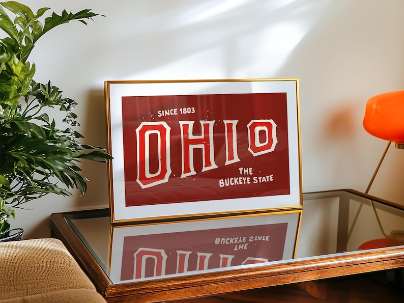 K&ouml;nnte beinhalten: Ein goldfarbener gerahmter Druck mit rotem Hintergrund und wei&szlig;em Text, der "Since 1803 OHIO The Buckeye State" lautet.
