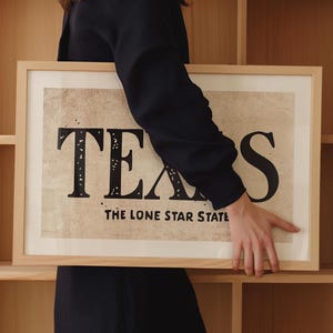 May include: A framed print with a vintage design featuring the word "TEXAS" in large, bold, black letters with a distressed look. The text "THE LONE STAR STATE" is printed below the word "TEXAS".