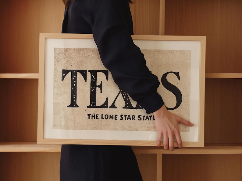 May include: A framed print with a vintage design featuring the word "TEXAS" in large, bold, black letters with a distressed look. The text "THE LONE STAR STATE" is printed below the word "TEXAS".