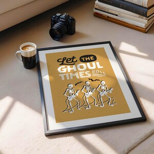 May include: Framed art print with the text "Let the Ghoul Times Roll" and illustrations of skeleton musicians playing guitars. The print has a mustard yellow background and a black frame. A black camera and a coffee mug are also visible.