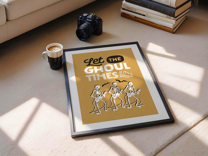 May include: Framed art print with the text "Let the Ghoul Times Roll" and illustrations of skeleton musicians playing guitars. The print has a mustard yellow background and a black frame. A black camera and a coffee mug are also visible.