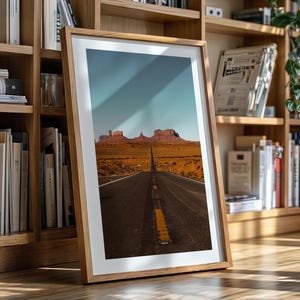 May include: A framed photograph of a long, straight road leading towards a mountain range in the distance. The road is paved and has a yellow dividing line. The mountains are brown and rocky. The sky is blue and cloudy.