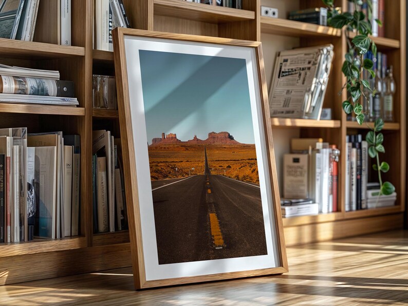May include: A framed photograph of a long, straight road leading towards a mountain range in the distance. The road is paved and has a yellow dividing line. The mountains are brown and rocky. The sky is blue and cloudy.