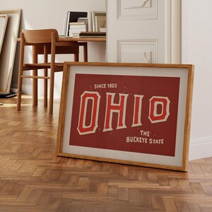 K&ouml;nnte beinhalten: Ein gerahmter Druck mit rotem Hintergrund und wei&szlig;em Text, der "OHIO SINCE 1803 THE BUCKEYE STATE" lautet. Der Text ist in einer Schriftart im Vintage-Stil.