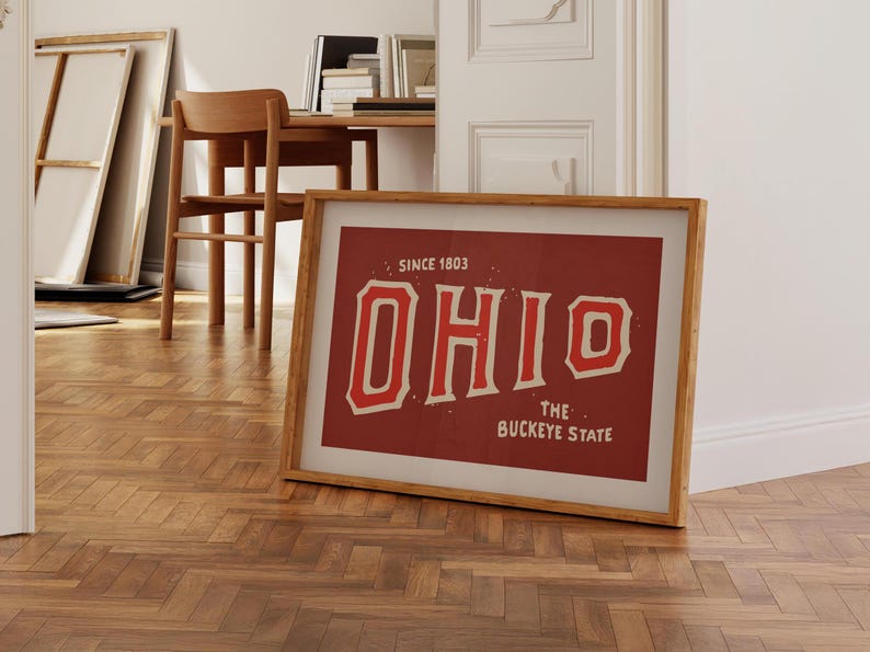 K&ouml;nnte beinhalten: Ein gerahmter Druck mit rotem Hintergrund und wei&szlig;em Text, der "OHIO SINCE 1803 THE BUCKEYE STATE" lautet. Der Text ist in einer Schriftart im Vintage-Stil.