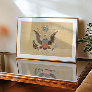 K&ouml;nnte beinhalten: Ein gerahmter Druck mit dem Gro&szlig;en Siegel der Vereinigten Staaten, mit einem goldenen Rahmen. Das Kunstwerk zeigt den amerikanischen Wei&szlig;kopfseeadler, der einen Olivenzweig und Pfeile h&auml;lt, mit einem Schild und Sternen dar&uuml;ber. Der Druck befindet sich auf einem Glastisch.