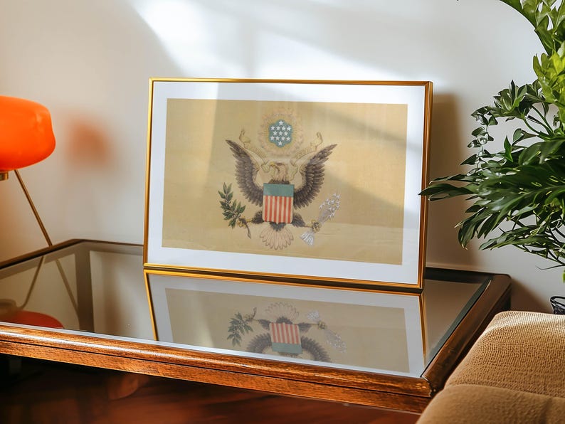 K&ouml;nnte beinhalten: Ein gerahmter Druck mit dem Gro&szlig;en Siegel der Vereinigten Staaten, mit einem goldenen Rahmen. Das Kunstwerk zeigt den amerikanischen Wei&szlig;kopfseeadler, der einen Olivenzweig und Pfeile h&auml;lt, mit einem Schild und Sternen dar&uuml;ber. Der Druck befindet sich auf einem Glastisch.