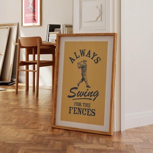 May include: A framed baseball-themed print with the text "ALWAYS Swing for the FENCES" in navy blue on a tan background. The image features a baseball player in action. The frame is a light brown color.