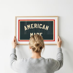 May include: A framed sign with the words "AMERICAN MADE" in white lettering on a dark teal background, framed with a red border. The frame is a light wood colour. A person is holding the framed sign.