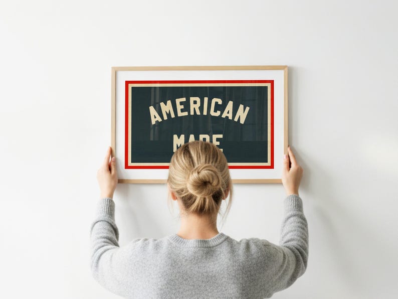May include: A framed sign with the words "AMERICAN MADE" in white lettering on a dark teal background, framed with a red border. The frame is a light wood colour. A person is holding the framed sign.
