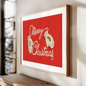 May include: A framed Christmas print with a red background. The artwork features the words "Merry Christmas" in cursive script, along with two cream-colored mittens decorated with holly. The frame is a light brown color.