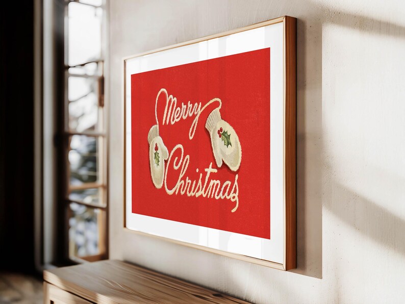 May include: A framed Christmas print with a red background. The artwork features the words "Merry Christmas" in cursive script, along with two cream-colored mittens decorated with holly. The frame is a light brown color.