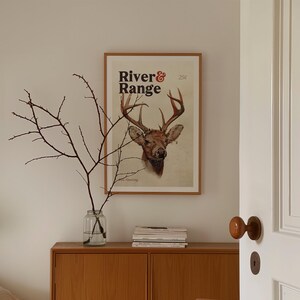 May include: A framed print with a vintage illustration of a deer head with antlers. The print has the text "River & Range" in a bold, black font.