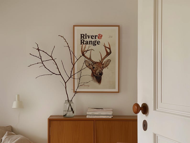 May include: A framed print with a vintage illustration of a deer head with antlers. The print has the text "River & Range" in a bold, black font.