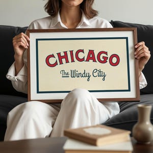 Op de afbeelding: Een ingelijste print met het woord "CHICAGO" in rode, gebogen letters en "The Windy City" in een scriptlettertype. De print heeft een cr&egrave;mekleurige achtergrond en een donkerblauwe rand, in een bruine lijst.
