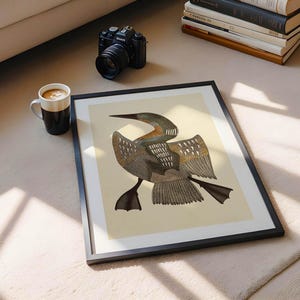 May include: A framed art print featuring a detailed illustration of a bird with outstretched wings, in shades of brown, gray, and beige. The artwork is set against a cream background, within a black frame. A coffee mug, camera, and books are also visible.