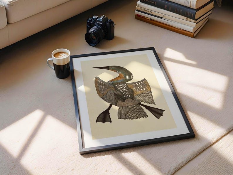 May include: A framed art print featuring a detailed illustration of a bird with outstretched wings, in shades of brown, gray, and beige. The artwork is set against a cream background, within a black frame. A coffee mug, camera, and books are also visible.