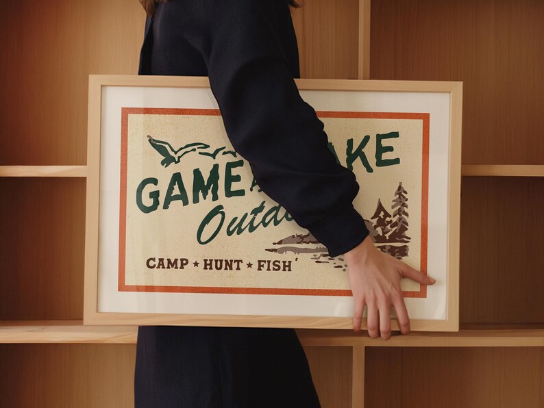 Op de afbeelding: Een ingelijst drukwerk met een vintage ontwerp met de tekst "Game Lake Outdoors" en de woorden "Camp, Hunt, Fish" in een kleiner lettertype. De print heeft een cr&egrave;mekleurige achtergrond met groene en bruine accenten.