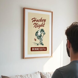 Puede incluir: P&oacute;ster enmarcado con un dise&ntilde;o vintage. El p&oacute;ster presenta las palabras "Hockey Night" en una fuente de escritura sobre una ilustraci&oacute;n de un jugador de hockey. Debajo de la ilustraci&oacute;n, el texto "IN MINNESOTA" se muestra en un recuadro.